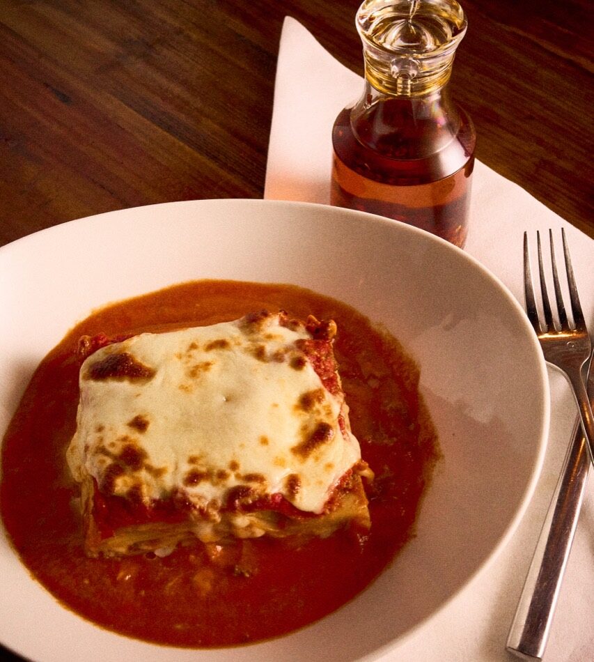Slice of Mom's lasagna in tomato sauce with a bottle of chili oil on the table